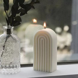 A tall, white pillar candle with a ribbed, arched design. The candle is placed on a windowsill next to a glass vase filled with dark leaves.