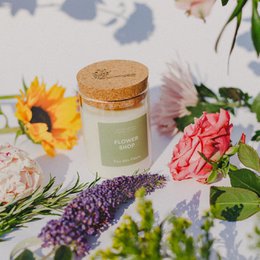 Floral scented candle in a glass jar with a cork lid. The candle is surrounded by fresh flowers, including sunflowers and pink roses. The candle is labeled "Flower Shop" and comes in various sizes.