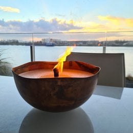 A round, tall copper-colored candle in a bowl-shaped container, with a flame burning. The candle is placed on a table with a scenic view of a body of water and cityscape in the background.