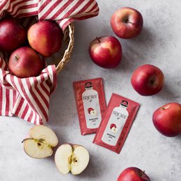 Red apples in a woven basket, accompanied by red and white striped fabric and apple juice cartons. The apples appear to be fresh and ripe, with a vibrant red color.