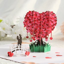 A 3D pop-up card featuring a heart-shaped tree made of red paper hearts, with a couple standing in front of it. The card is designed for Valentine's Day.