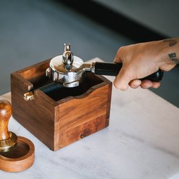 A wooden knock box with a metal lever for tamping coffee grounds. The box is made of dark brown wood and has a simple, rustic design.