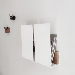 Wall-mounted magazine rack in black, white, grey, and green colors. Rectangular shape with open compartments to store magazines and other reading materials.