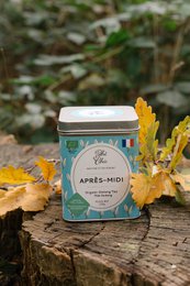 Organic tea in a teal tin with a floral label. The tea is served in a white teapot with a woman in a yellow sweater pouring the tea.