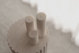 Tall, cylindrical white pillar candles made of wax. The candles have a smooth, uniform texture and are arranged in a group.