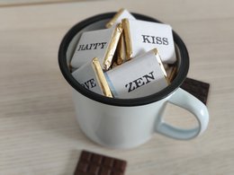 A mug filled with chocolates labeled "Happy" and "Kiss" in a black and white color scheme, accompanied by pieces of chocolate.

The image shows a mug filled with chocolates that have the words "Happy" and "Kiss" printed on them. The chocolates are arranged in a circular pattern inside the mug, which has a black and white color scheme. Alongside the mug, there are a few pieces of chocolate visible.