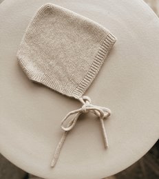Soft, cozy merino wool baby bonnet in a neutral sand color. Features a warm, knitted design with ties to secure a snug fit.