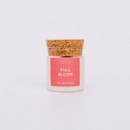 A tall, round white candle with a pink label that says "Full Bloom". The candle is surrounded by a gold textured material, likely a decorative lid or container.