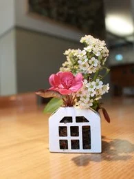 A white ceramic vase with a pink flower and small white flowers arranged inside. The vase has a simple, minimalist house-like shape.