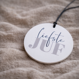  A round white keychain with the text "Lifestyle" printed on it in a cursive font, hanging on a black cord against a beige fabric background.
