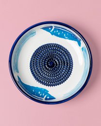  A round, shallow ceramic dish with a blue and white pattern featuring a central spiral design. The dish appears to be used for grating or shredding food items, as indicated by the hand holding a tomato and grating it in the second image.