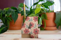  A floral patterned pencil holder with a wooden base and a cutout design. The holder is placed on a wooden surface with potted plants in the background.