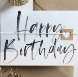 A folding birthday card with a blue envelope. The card features the text "Happy Birthday" in a handwritten style with a small heart symbol.