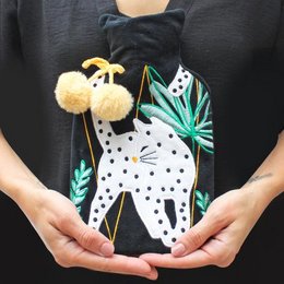 A black hot water bottle with a feline design, featuring a spotted cat-like pattern and pom-pom accents. This product is categorized as a sleep mask.