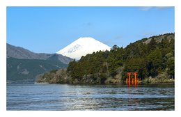 Panoramic landscape with a red Torii gate in the foreground and a snow-capped mountain in the background. The image depicts a serene lake surrounded by lush green forests.