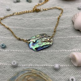 Iridescent paua/abalone shell pendant on a gold-toned chain. The shell has a natural, organic shape and displays a range of colors including green, blue, and purple.