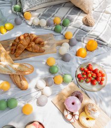 Outdoor string lights with colorful decorative balls, croissants, and fresh strawberries on a cozy blanket. The image suggests a relaxing, outdoor picnic or gathering with a variety of textures and colors.