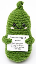  A green, crocheted pickle toy with a friendly face and a tag that reads "Emotional Support Pickle". The toy appears to be soft and plush, with a whimsical design.