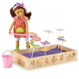 A colorful, whimsical garden-themed doll set with a young girl figure, accessories, and a wooden play box. The doll has long dark hair and is wearing a pink top and green pants. The set includes various garden-inspired elements like flowers, mushrooms, and a watering can.
