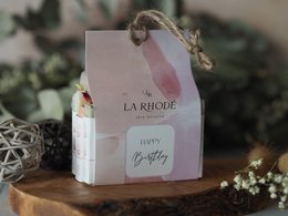 A pink and white gift box with the text "La Rhode" and "Happy Birthday" printed on it. The box is placed on a wooden surface with some greenery around it.