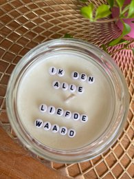  A round, tall candle with a rose quartz gemstone. The candle has a hidden message that reads "Ik ben alle liefde waard" which translates to "I am worthy of all love". The candle is scented with essential oils from DoTerra.