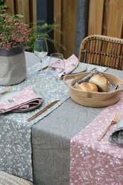 A handmade table runner in various colors, including grey, pink, sahara, and olive, measuring 40x150 cm. The runner is displayed on a rustic outdoor setting with a wicker chair, potted plants, and a basket of bread.