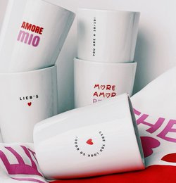  A white porcelain mug with a heart-shaped design and various romantic text messages printed on it, including "AMORE MIO", "LIEB'S", "BABE YOU LOOK SO GOOD!", "MORE AMOR POR FAVOR", and "YOU ARE A 10/10!". The mug is held in a hand with red nail polish.