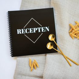  A black spiral-bound recipe book with the title "RECEPTEN" in white text, surrounded by golden utensils and ingredients on a beige background.