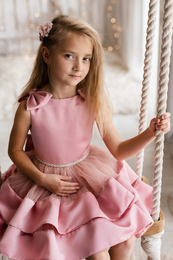 A pink, ruffled dress for girls with a bow detail. The dress has a sleeveless design and a full, layered skirt. It is suitable for formal or special occasions.
