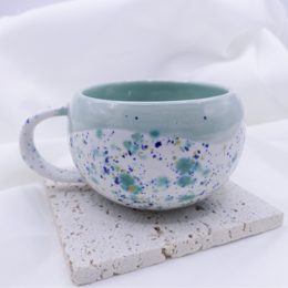 A ceramic mug featuring a winter forest design with green pine trees and white stars on a light background. The mug is being held in hands wrapped in a cozy, fuzzy blanket, creating a warm and cozy atmosphere.