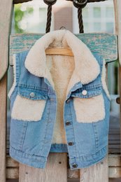 A denim vest with a cozy, sherpa-lined interior for kids. The vest features a hooded design and two front pockets.