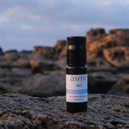  A small black glass bottle of essential oil labeled "ASMI" sits on a rocky surface. The bottle is accompanied by a white cylindrical container, also labeled "ASMI".