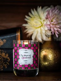 A round, tall pink and white scented candle with a floral pattern label. The candle is surrounded by pink and white flowers, creating a cozy and inviting atmosphere.