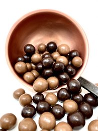 Assorted milk and dark chocolate balls in a wooden bowl. The chocolates come in a variety of sizes and shapes, including round and oval forms. The colors range from light brown to dark brown, indicating a mix of milk and dark chocolate.