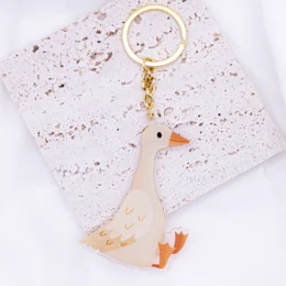 Acrylic keychain in the shape of a goose with a gold-colored ring. The goose has a white body, orange beak, and red accents.