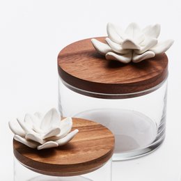Round glass storage jar with wooden lid and white ceramic flower decoration. Suitable for storing various items.