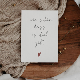 A simple, handwritten postcard with the text "we given days is such a gift" and a small red heart. The postcard is placed on a wooden surface with a beige fabric in the background.