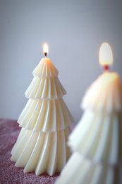 Tall, tiered white candle in the shape of a Christmas tree with a flame at the top. The candle has a layered, textured appearance.