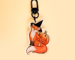  Decorative keychain featuring an orange fox design with a pumpkin. The fox is sitting and has a whimsical, cartoon-like appearance.