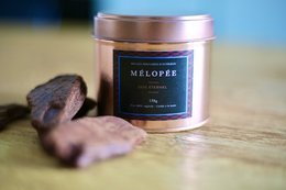 Tall, round candle with a dark label featuring the text "MELOPEE Eternal Oud". The candle appears to be made of a dark-colored wax and is placed on a wooden surface.