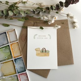 A greeting card featuring a watercolor illustration of a moses basket, surrounded by greenery and floral elements. The card has a simple, natural aesthetic.