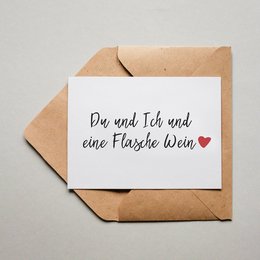 A greeting card with the text "Du und Ich und eine Flasche Wein ❤️" (You and I and a bottle of wine ❤️) written on it, enclosed in a brown envelope.