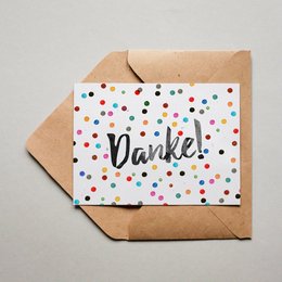 Colorful thank you card with the German word "Danke" (meaning "Thank you") written in a stylized font, surrounded by a pattern of multicolored dots or confetti. The card is placed on a brown paper envelope.