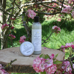 Wooly Wash 270ml laundry detergent in cedar wood, earthy essence, patchouli passion, and perfectly pure (no perfume) scents. Bottle and tin container shown among pink blossoms.