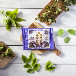 A single protein cookie in a purple and yellow wrapper, featuring floral and geometric designs. The cookie appears to be a healthy snack option.