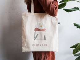  Fairtrade tote bag made of natural cotton material, featuring a simple text design "One day at a time" printed on the front. The bag has a minimalist and eco-friendly style.