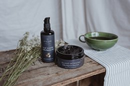 Black gel body product in glass bottle and jar, with green ceramic bowl on wooden table.