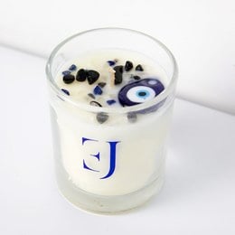 Scented candle with evil eye pendant and crystals, round glass container, white wax with black and blue accents.