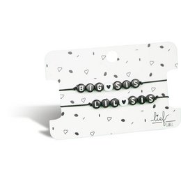 A set of two black and white charm bracelets, one labeled "Big Sis" and the other "Lil Sis", presented on a patterned background with hearts.