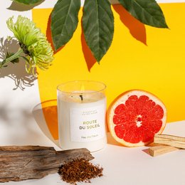  A tall, round white candle with a cork lid, surrounded by autumn leaves and a grapefruit slice. The candle is labeled "Route du Soleil", indicating a scented candle product.